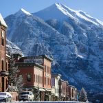 Snow-covered town in the Rocky Mountains showcasing historic buildings and parked vehicles under a clear blue sky.