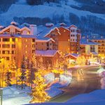 Ski resort at dusk with holiday lights, snow-covered ground, and pine trees, near Rocky Mountain real estate.