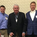Three men in formal attire discussing real estate trends at a networking event, with a neutral textured wall background.
