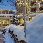Snow-covered landscape with warm-lit buildings, holiday lights, and a tree near Rocky Mountain real estate properties.