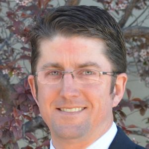 Middle-aged man in a suit at Rocky Mountain Commercial Real Estate, surrounded by a blurred natural background.
