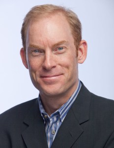 Professional man in a suit jacket with light brown hair and blue eyes against a neutral background, smiling warmly.