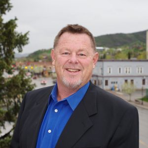 Smiling man in a black blazer and blue shirt, representing Rocky Mountain Commercial Real Estate in an urban setting.