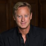 Man in a black suit jacket with a dark wooden backdrop, representing Rocky Mountain Commercial Real Estate professionalism.