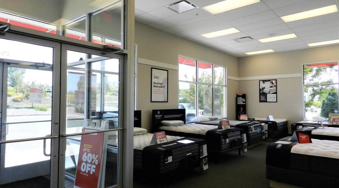 Interior of a commercial mattress showroom featuring multiple beds on display, large windows allowing natural light, promotional signage indicating "UP TO 60% OFF," and a wall poster addressing sleep issues like snoring and back pain.