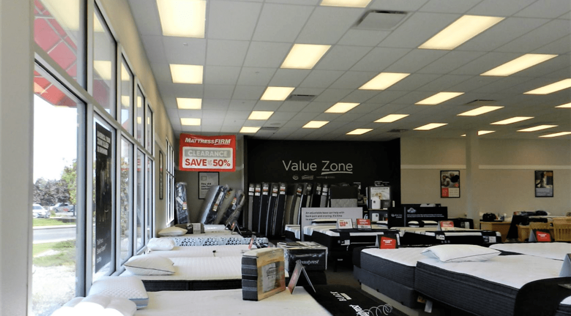 Interior of a mattress showroom featuring various displayed mattresses, a "Mattress Firm" clearance sign stating "SAVE 50%", and a section labeled "Value Zone", emphasizing the commercial retail aspect of the property for sale at 3501 Wolverine Dr, Montrose, Colorado.