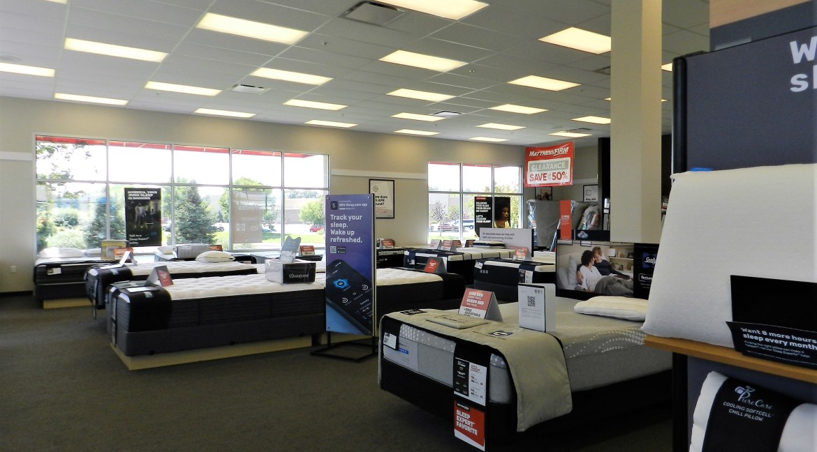 Showroom displaying various mattresses and bedding products with promotional signage, including clearance offers and advertisements for sleep tracking technology, in a well-lit retail space.