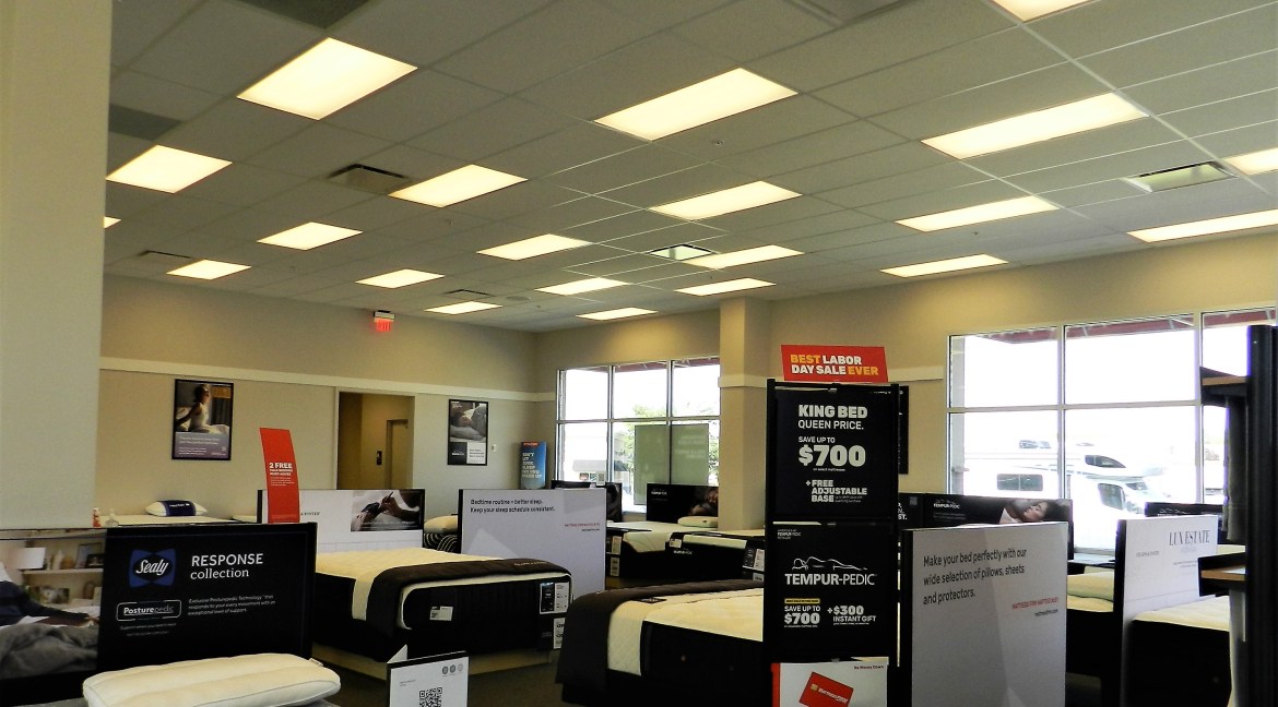 Showroom interior featuring various mattress displays, promotional signage for Labor Day sale, and large windows allowing natural light, highlighting a commercial retail space for bedding products.