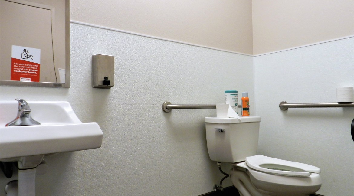 Restroom interior featuring a white sink, toilet, hand sanitizer dispenser, and toilet paper, emphasizing cleanliness and accessibility.