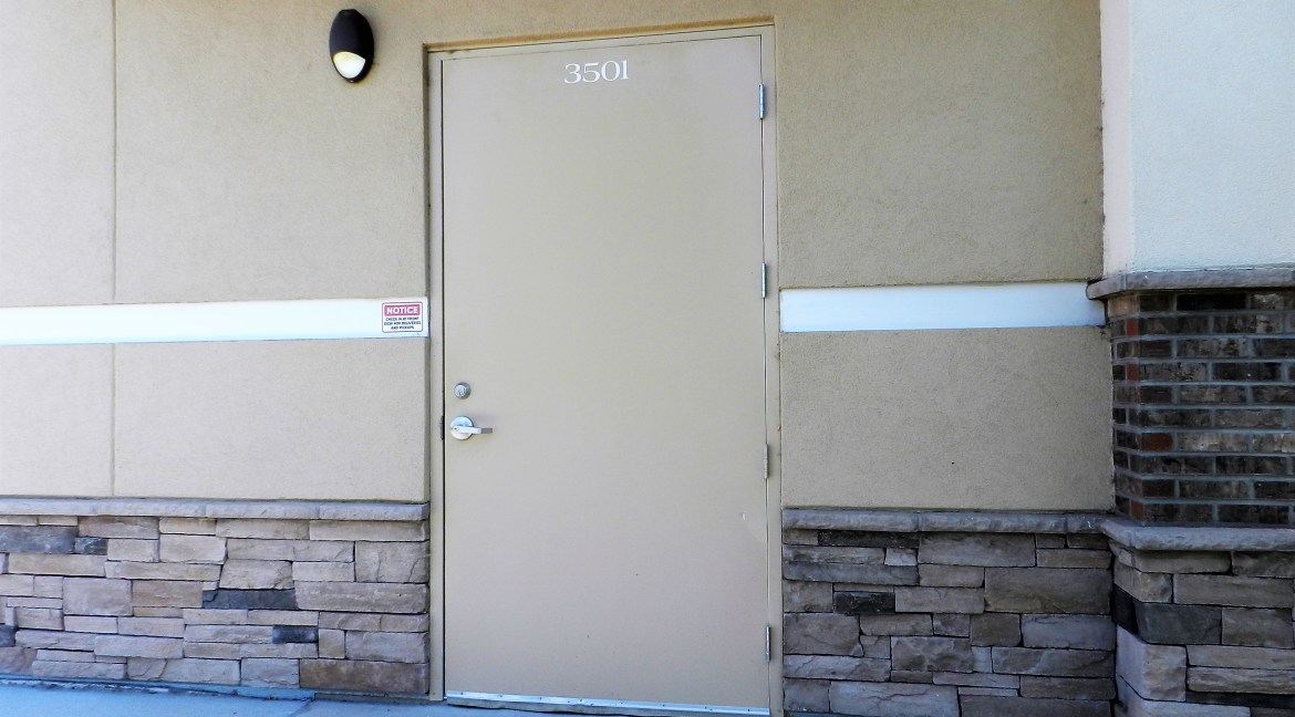 Commercial building entrance at 3501 Wolverine Dr, featuring a beige door with the number "3501," stone base, and notice sign, part of a retail/office property for sale in Montrose, Colorado.