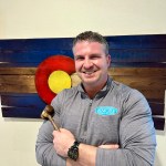 David Porter, Broker/Owner of Rustic Realty, smiling with a gavel in front of a Colorado flag backdrop, representing leadership in real estate in Colorado Springs.