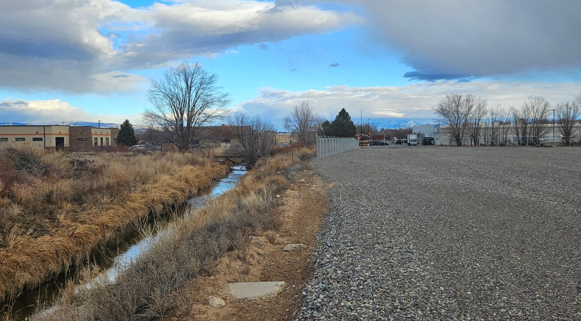4.34-acre commercial land for sale at 65 Circle Dr., Delta, Colorado, featuring a gravel lot, adjacent creek, and surrounding commercial buildings.