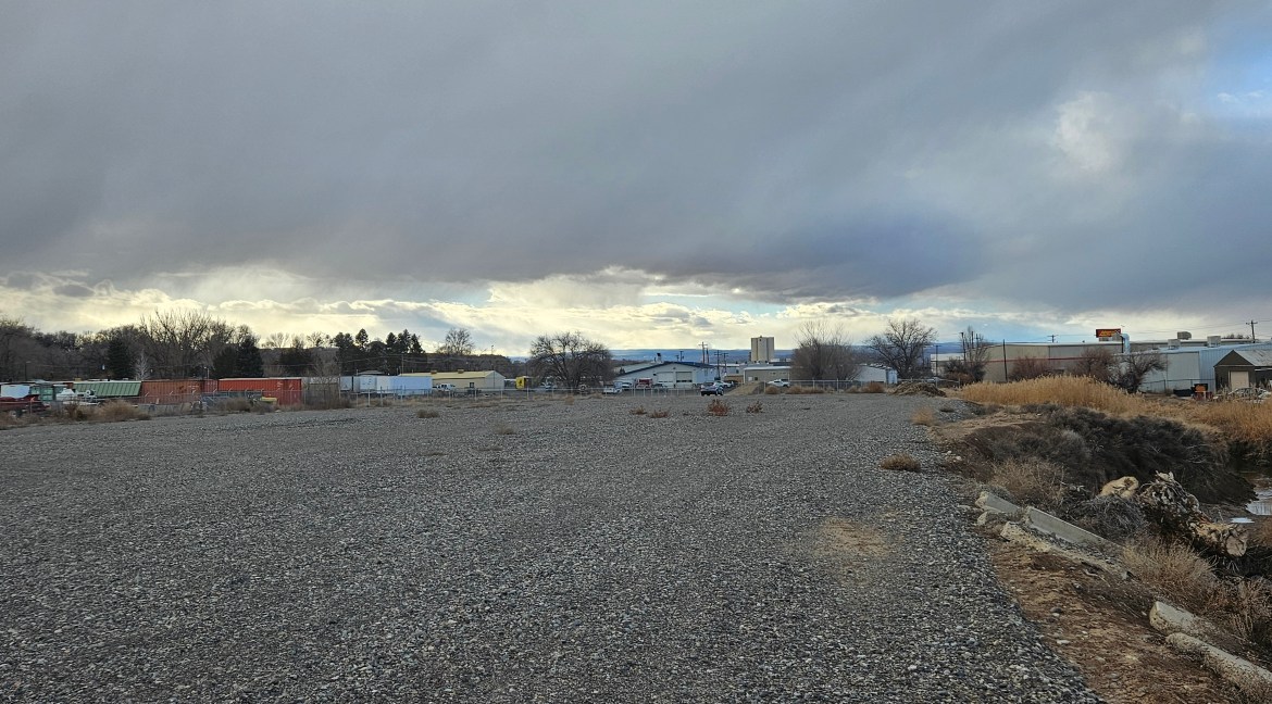 4.34 acres of vacant land with gravel surface and commercial buildings in the background, located near Highway 92 in Delta, Colorado, suitable for B-3 zoning development.