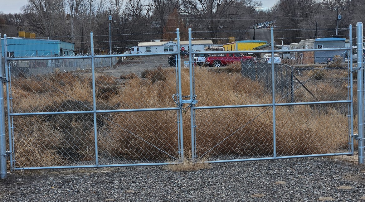 Fenced entrance to a commercial land parcel at 65 Circle Dr., Delta, Colorado, with dry grass and surrounding structures visible.