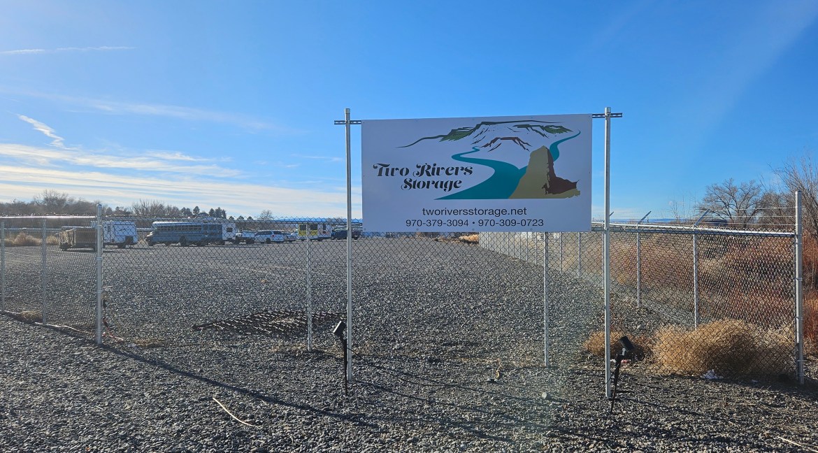 Sign for Two Rivers Storage with contact information, showcasing a gravel lot and storage area in Delta, Colorado, near commercial developments.