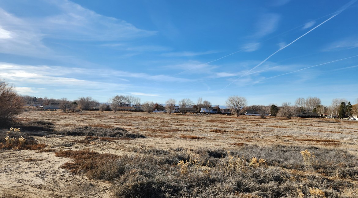 14.84-acre residential development land in Delta, Colorado, featuring open terrain, nearby trees, and distant homes under a clear blue sky, highlighting investment potential and proximity to commercial hubs.