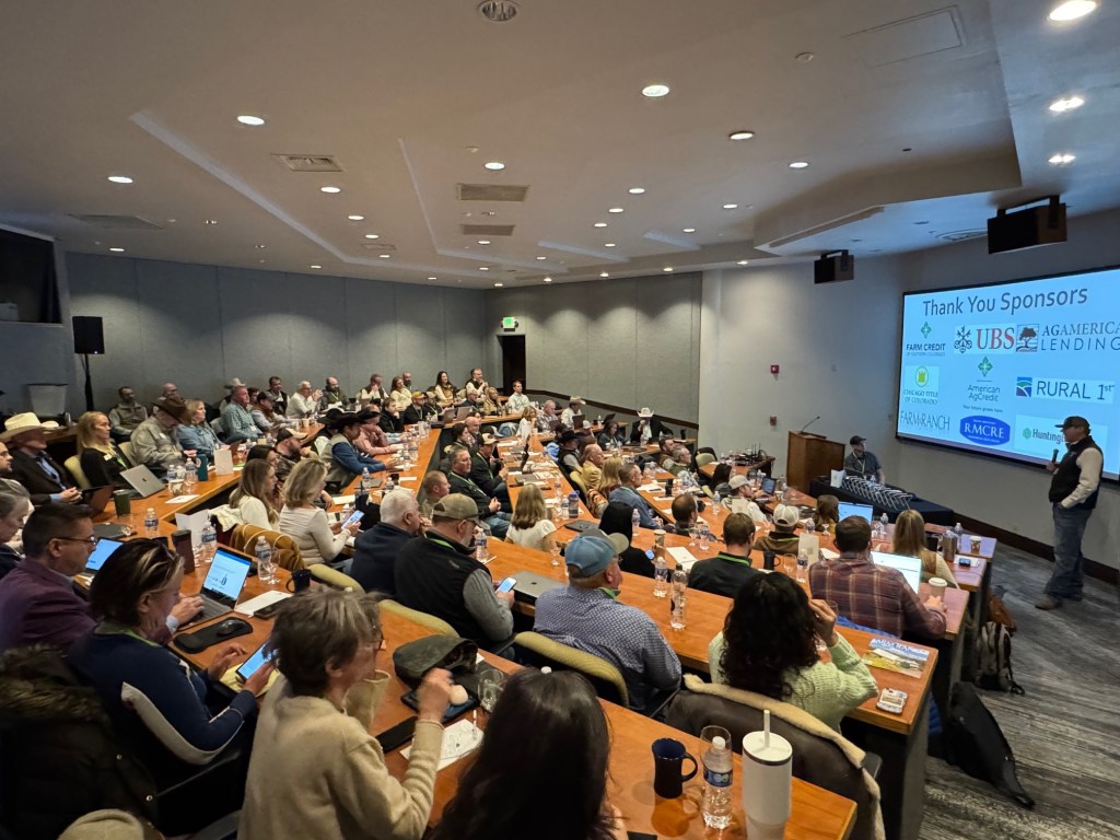 Audience at REALTORS® Land Institute seminar with RMCRE sponsor logos on screen and presenter in professional setting.