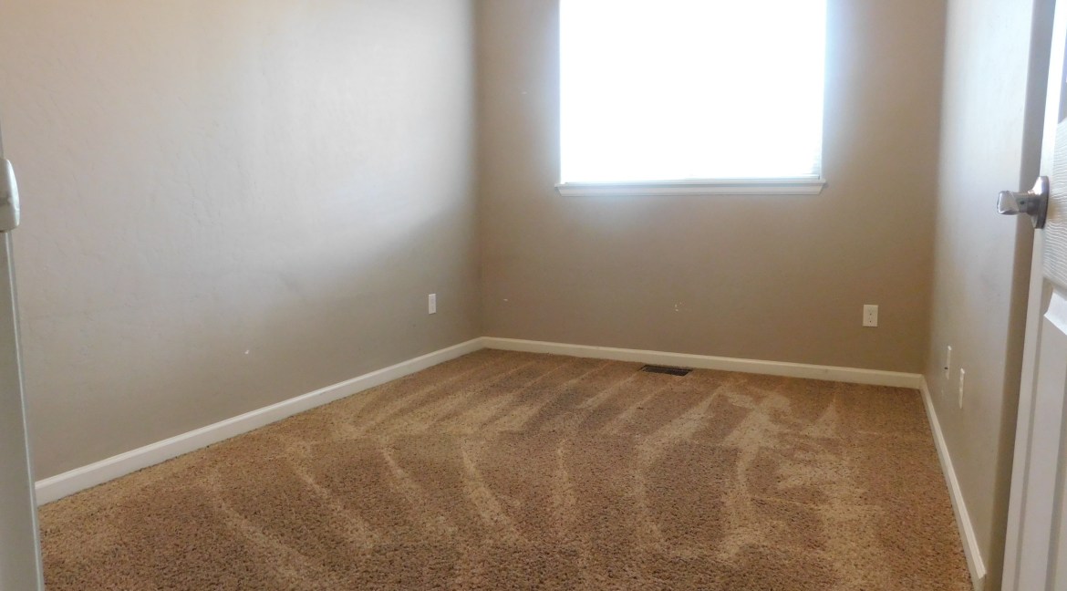 Empty bedroom with beige walls, carpeted floor, and a window allowing natural light, showcasing potential for customization in a modern townhome at 1534 Poplar Drive, Grand Junction, Colorado.