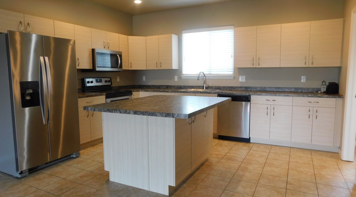 Modern kitchen with stainless steel appliances, including a refrigerator, microwave, and dishwasher, featuring light wood cabinets and a large island, designed for open-concept living in contemporary townhome units at 1534 Poplar Drive, Grand Junction, CO.