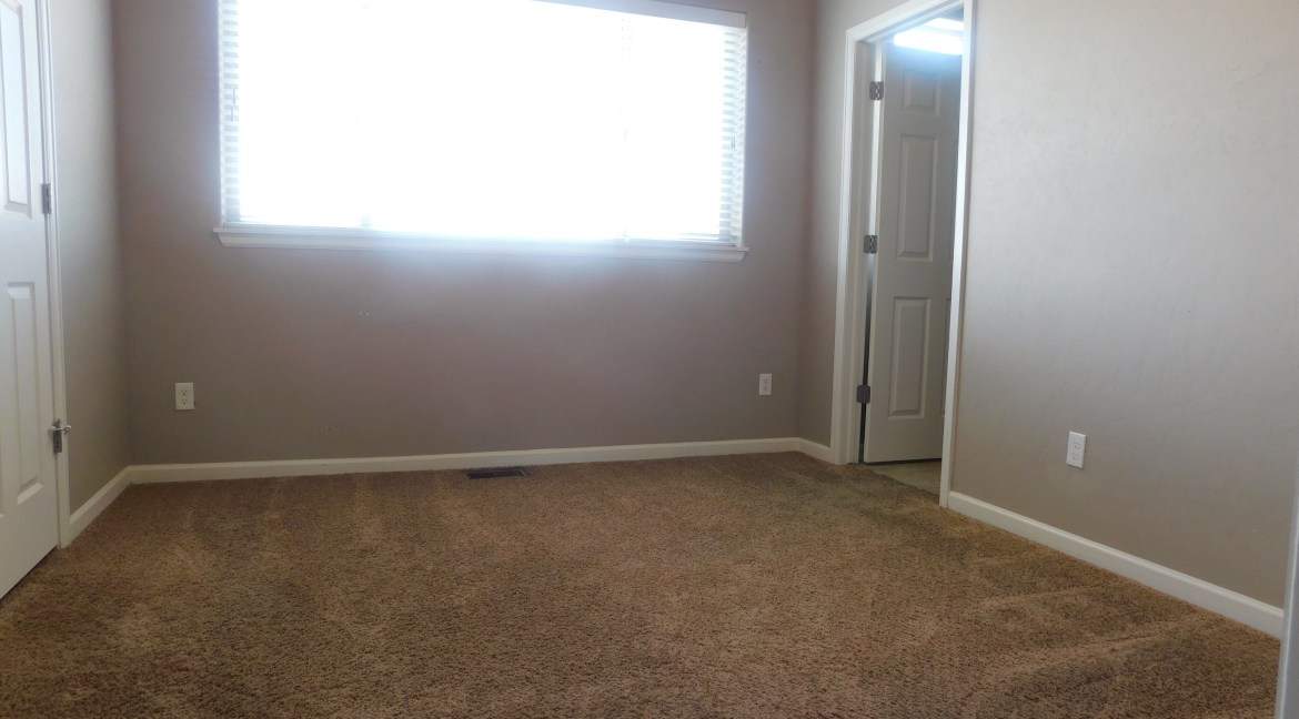 Interior view of a modern bedroom with carpet flooring, a large window allowing natural light, and two doors leading to adjacent rooms, emphasizing spaciousness in the townhome units at 1534 Poplar Drive, Grand Junction, CO.