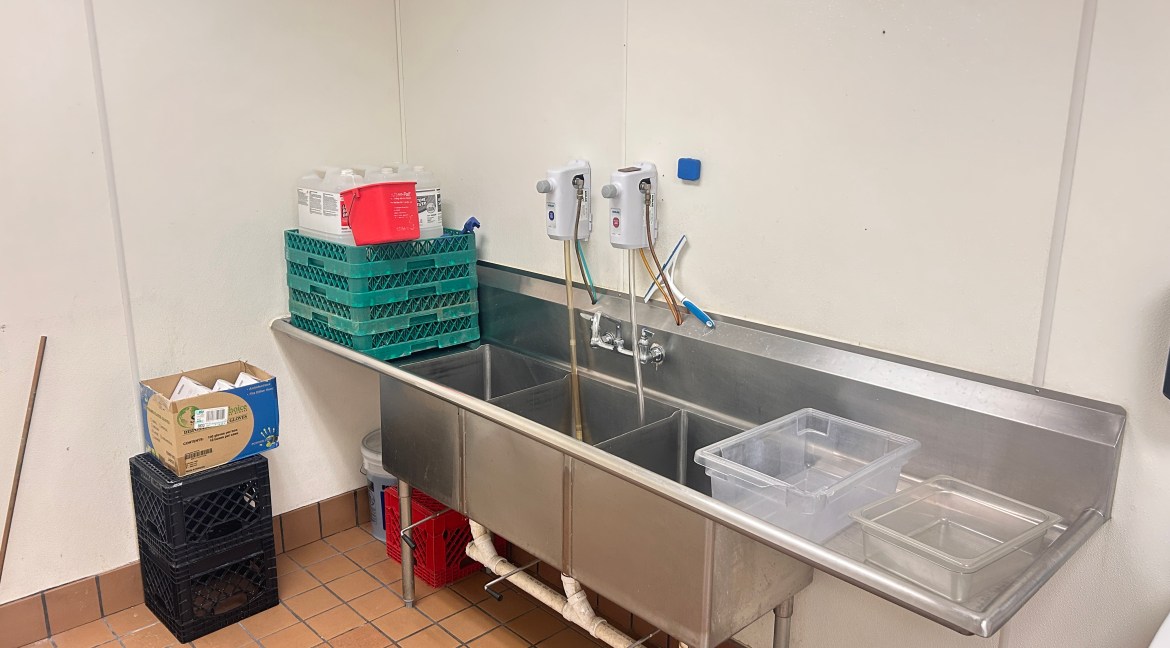 Commercial kitchen sink area with stainless steel three-compartment sink, cleaning supplies, and storage crates, highlighting features for food preparation and sanitation in a leased kitchen space at 350 Stafford Lane, Delta, Colorado.