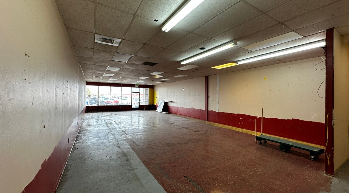 Retail space interior at Coronado Plaza Shopping Center, featuring vacant area with red and white walls, overhead lighting, and large windows offering visibility to the outside.