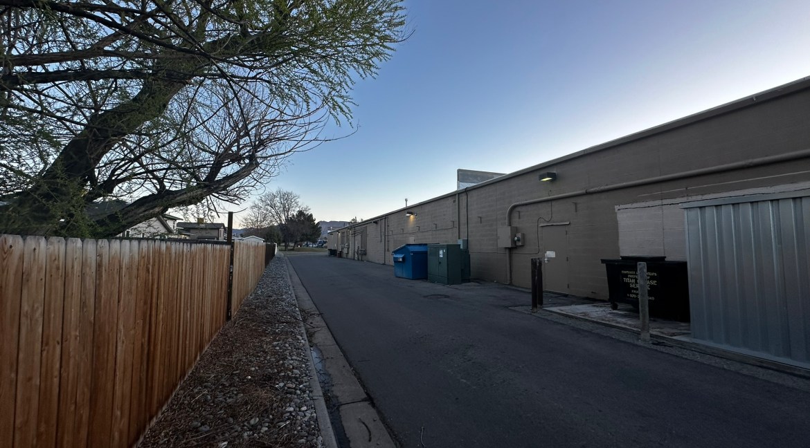 Retail unit access road at Coronado Plaza Shopping Center, Clifton, Colorado, featuring a wooden fence, dumpsters, and adjacent buildings.